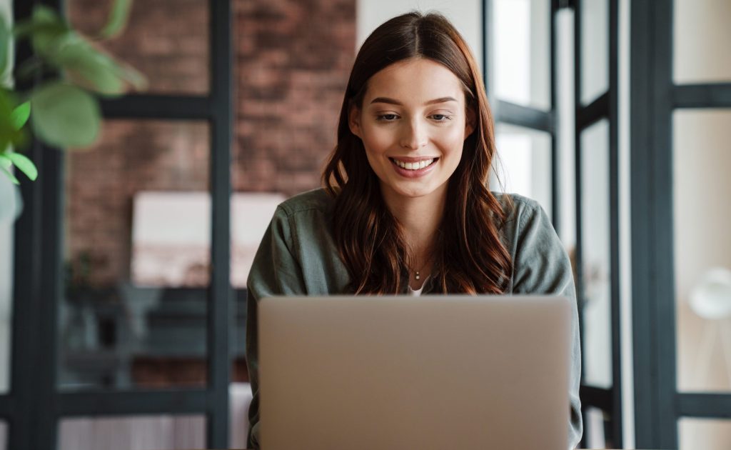 Beautiful smiling woman working with laptop while sitting at table indoors to get your real estate license online